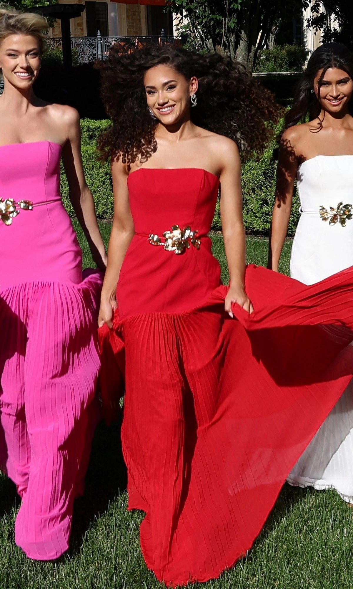 Three women walk on the grass, each wearing elegant strapless formal gowns in pink, red (Jovani 47000), and white, all featuring a gold floral waist detail. Trees and a building can be seen in the background.